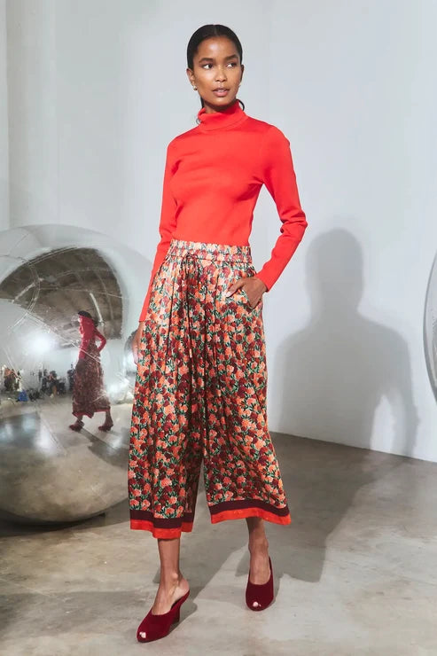 A model stands in a studio wearing a bright red mock-neck long-sleeve top, high-waisted wide-leg cropped pants with a red, orange, and green floral print, and red pointed-toe mule heels.