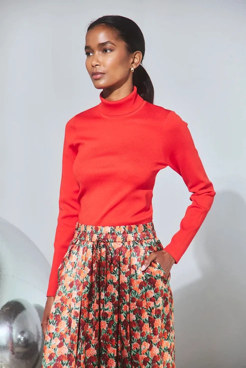 A woman modeling a bright red long-sleeve mock neck top paired with a floral print skirt, standing against a light gray studio background.