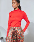 A woman modeling a bright red long-sleeve mock neck top paired with a floral print skirt, standing against a light gray studio background.