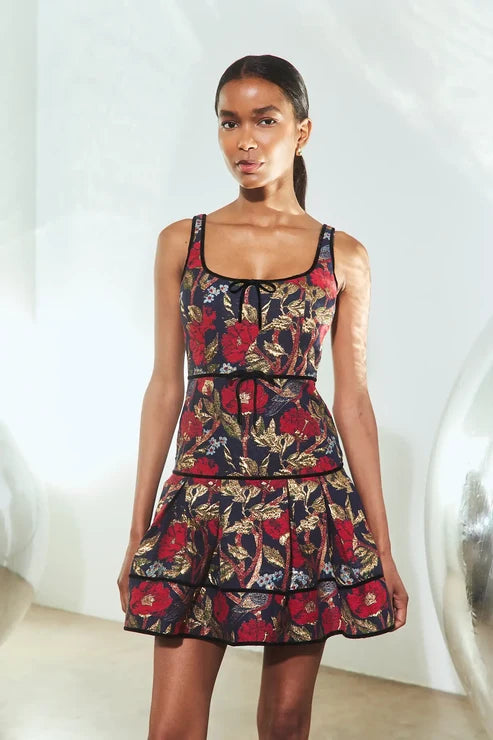 A woman modeling a sleeveless tiered mini dress with a dark navy background and a red, gold, and green floral print, standing in a brightly lit studio.