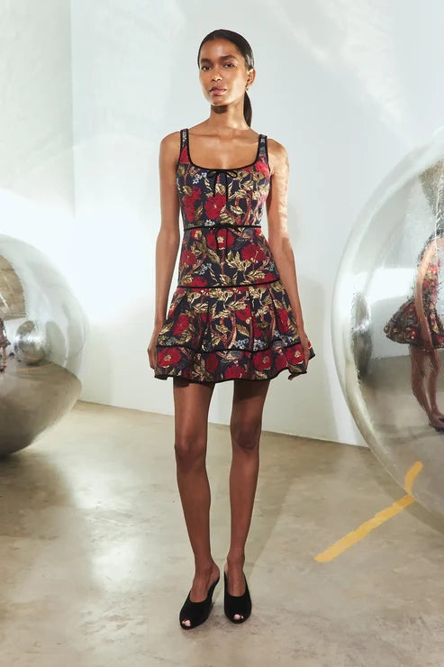 A model stands in a minimalist studio wearing a sleeveless mini dress with a dark floral print and black peep-toe heels. The background features white walls and large reflective glass spheres.