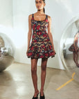 A model stands in a minimalist studio wearing a sleeveless mini dress with a dark floral print and black peep-toe heels. The background features white walls and large reflective glass spheres.