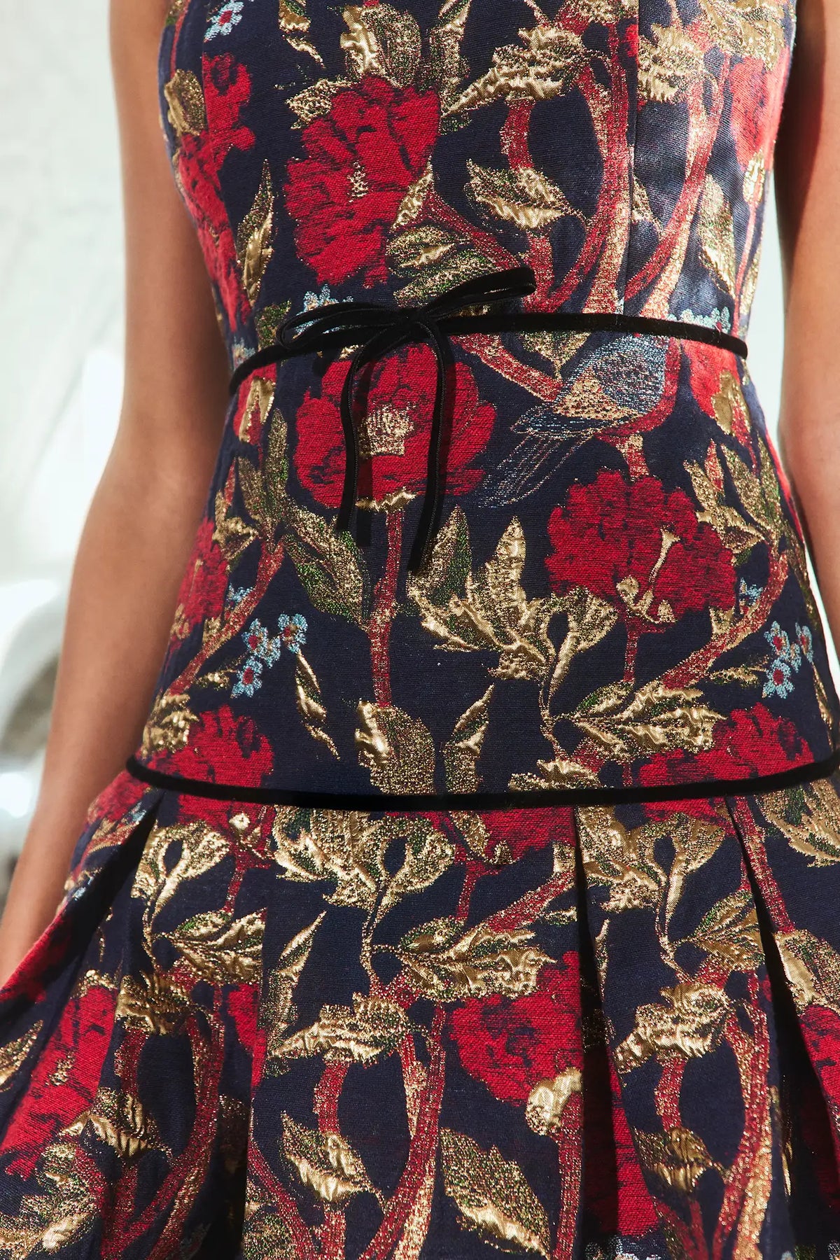 Close-up of a sleeveless dress with a dark navy background and an ornate floral pattern in metallic gold and deep red. The bodice is cinched at the waist with a thin black ribbon tie.