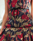 Close-up of a sleeveless dress with a dark navy background and an ornate floral pattern in metallic gold and deep red. The bodice is cinched at the waist with a thin black ribbon tie.