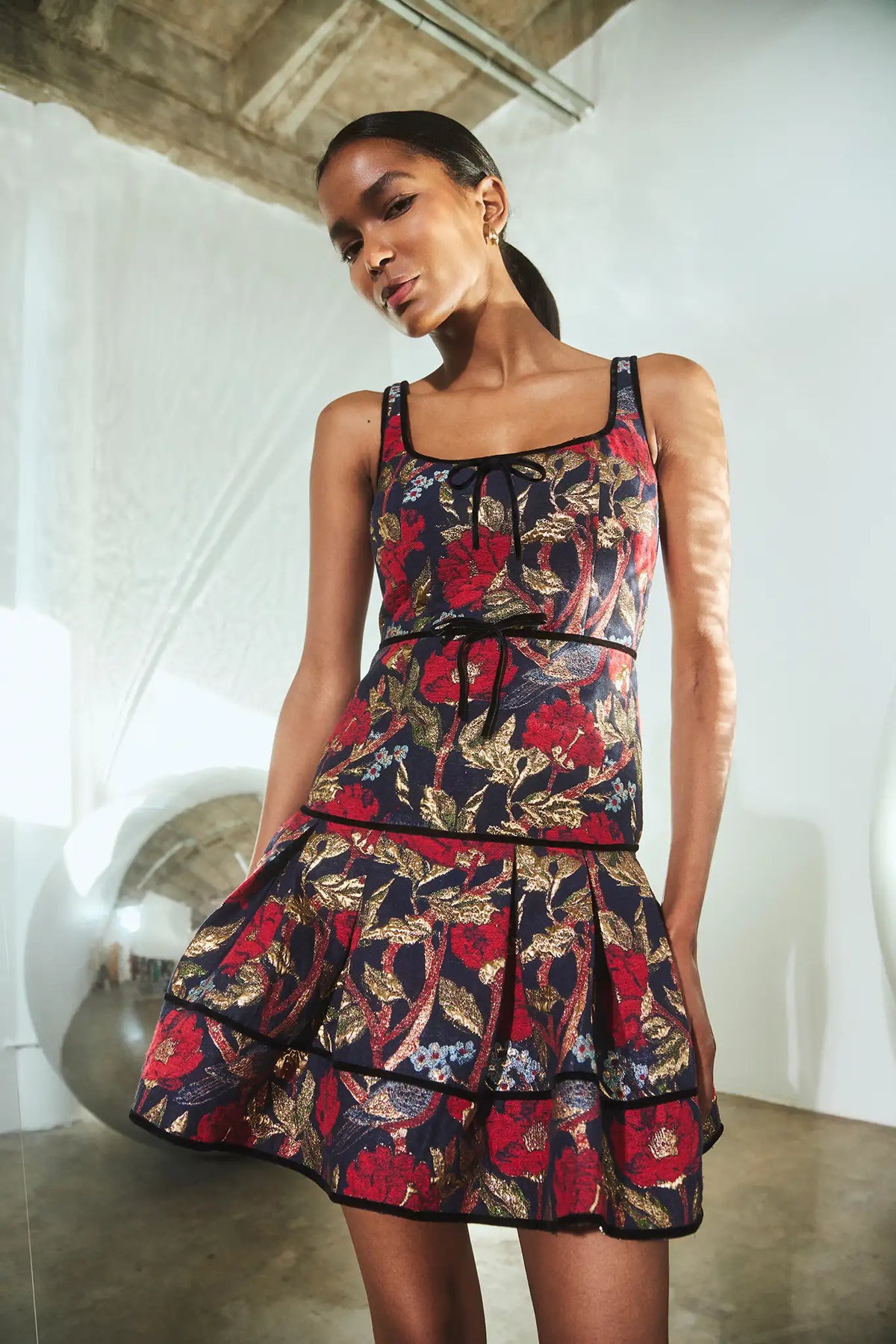 A model wearing a sleeveless tiered mini dress with a red, gold, and black floral print, standing in a modern white-walled studio with concrete floors.