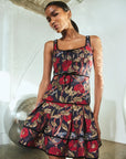 A model wearing a sleeveless tiered mini dress with a red, gold, and black floral print, standing in a modern white-walled studio with concrete floors.