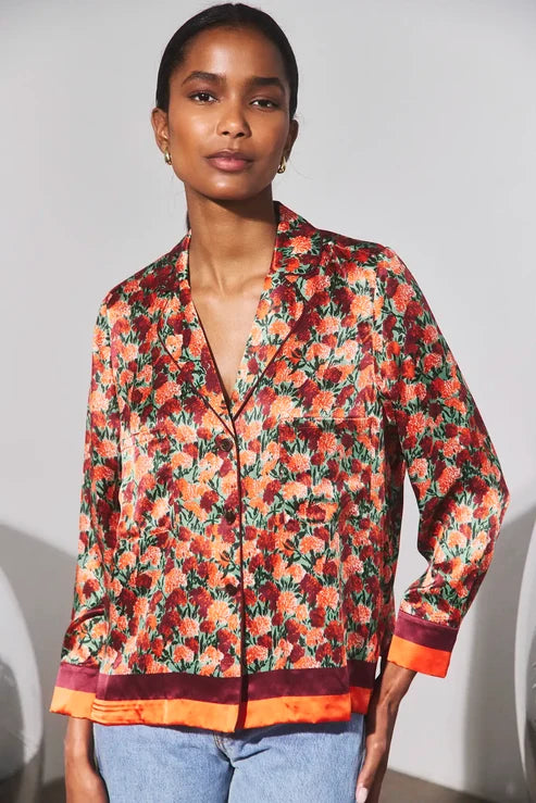 A woman wearing a long-sleeve satin-like button-front shirt jacket with an allover floral print in red, orange, green, and black, featuring contrasting orange and burgundy trim at the hem and cuffs, paired with light blue jeans.