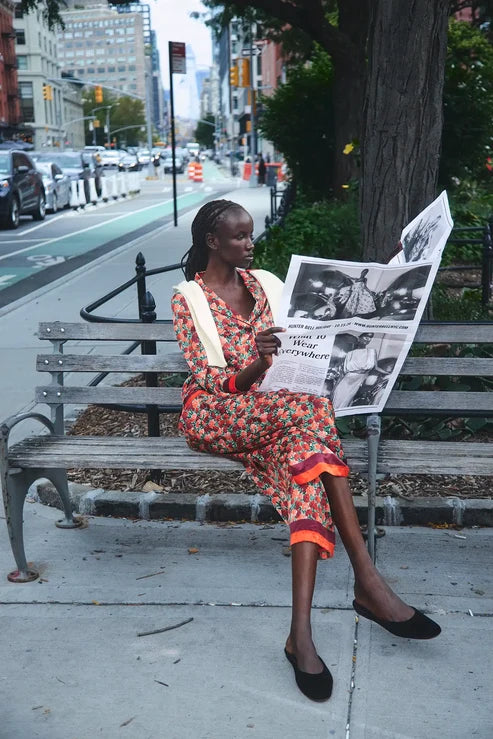 A woman sits on a city park bench reading a newspaper, wearing a colorful floral print dress with orange and yellow accents. The background shows a busy city street with cars, buildings, and trees.
