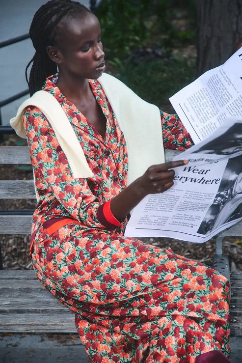 A woman sits on outdoor wooden steps holding a newspaper, wearing a long-sleeve red and orange floral dress with a cream shawl and red wristbands.