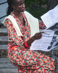 A woman sits on outdoor wooden steps holding a newspaper, wearing a long-sleeve red and orange floral dress with a cream shawl and red wristbands.