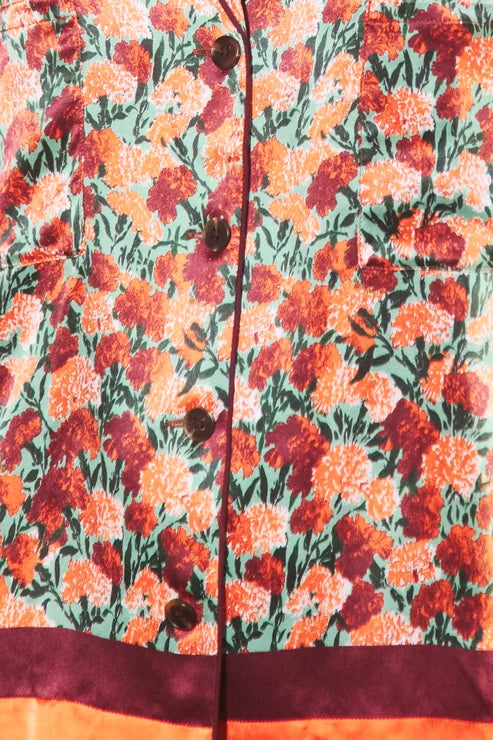Close-up of a floral-patterned garment, likely a jacket or coat, featuring orange, red, and yellow flowers with green leaves on a light background, with dark buttons and a dark burgundy border at the bottom.