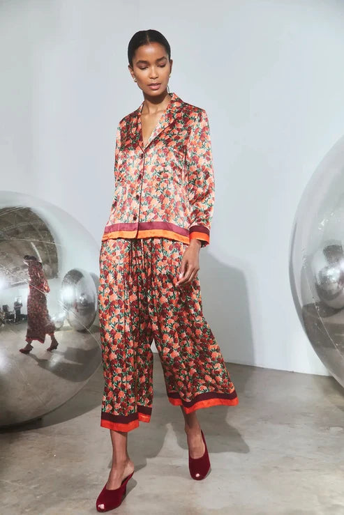 A model walks in a minimalist white studio wearing a matching two-piece pajama set with a dense red, orange, and green floral print, featuring a button-up shirt and wide-leg pants with contrasting orange-red trim, paired with dark red mule heels.