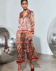 A model walks in a minimalist white studio wearing a matching two-piece pajama set with a dense red, orange, and green floral print, featuring a button-up shirt and wide-leg pants with contrasting orange-red trim, paired with dark red mule heels.