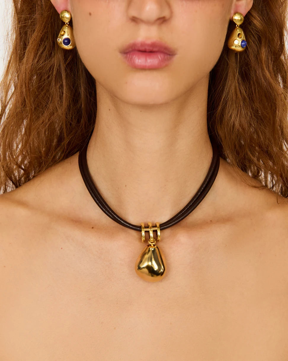 A close-up of a model wearing a black cord choker necklace with a shiny gold teardrop pendant, paired with matching gold and blue drop earrings.
