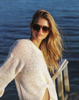 A woman with long blonde hair wearing a chunky knit white sweater and sunglasses, leaning on a wooden dock with a blue body of water in the background.