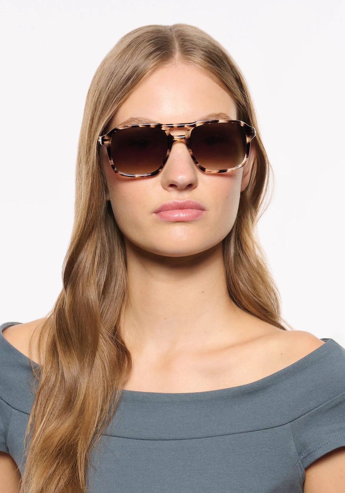 A woman with long light brown hair wearing oversized square tortoiseshell sunglasses with dark brown gradient lenses, set against a plain white background.