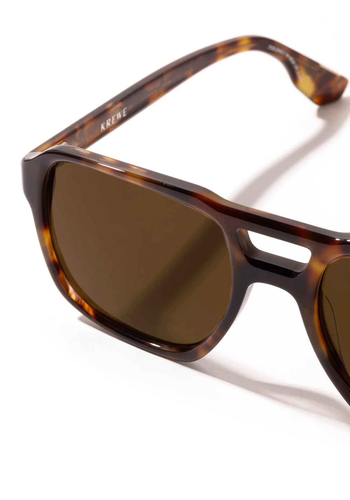 Close-up of a pair of sunglasses with a tortoiseshell acetate frame and dark brown lenses, showing the brand name Krewe on the inner temple arm, set against a clean white background.