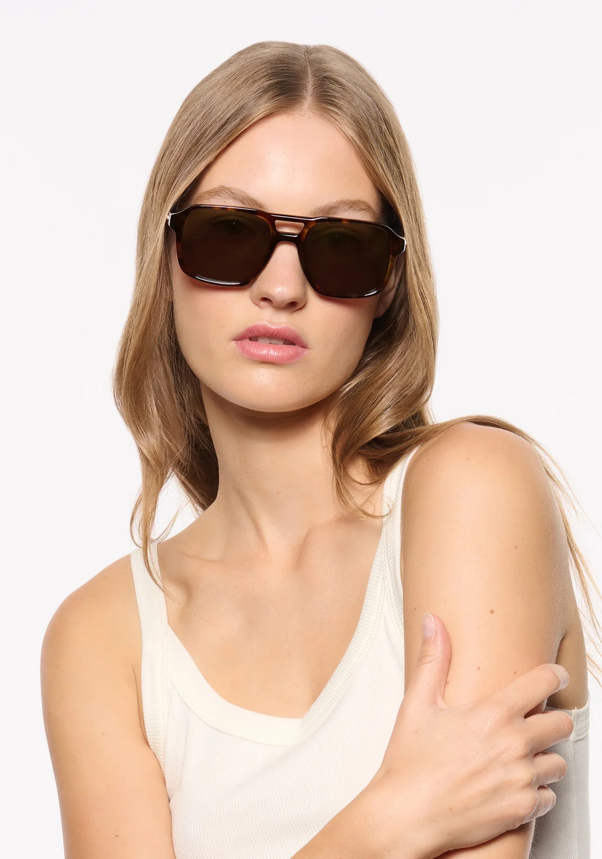 A woman with blonde hair wearing dark brown aviator-style sunglasses and a white tank top, photographed against a clean white background.