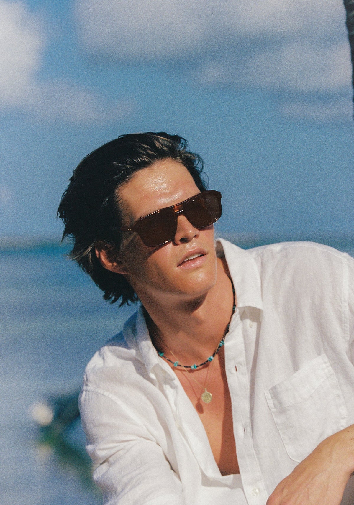 A man wearing dark brown aviator sunglasses, a white button-down shirt, and layered necklaces, photographed outdoors against a bright blue sky and ocean background.
