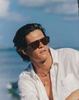 A man wearing dark brown aviator sunglasses, a white button-down shirt, and layered necklaces, photographed outdoors against a bright blue sky and ocean background.