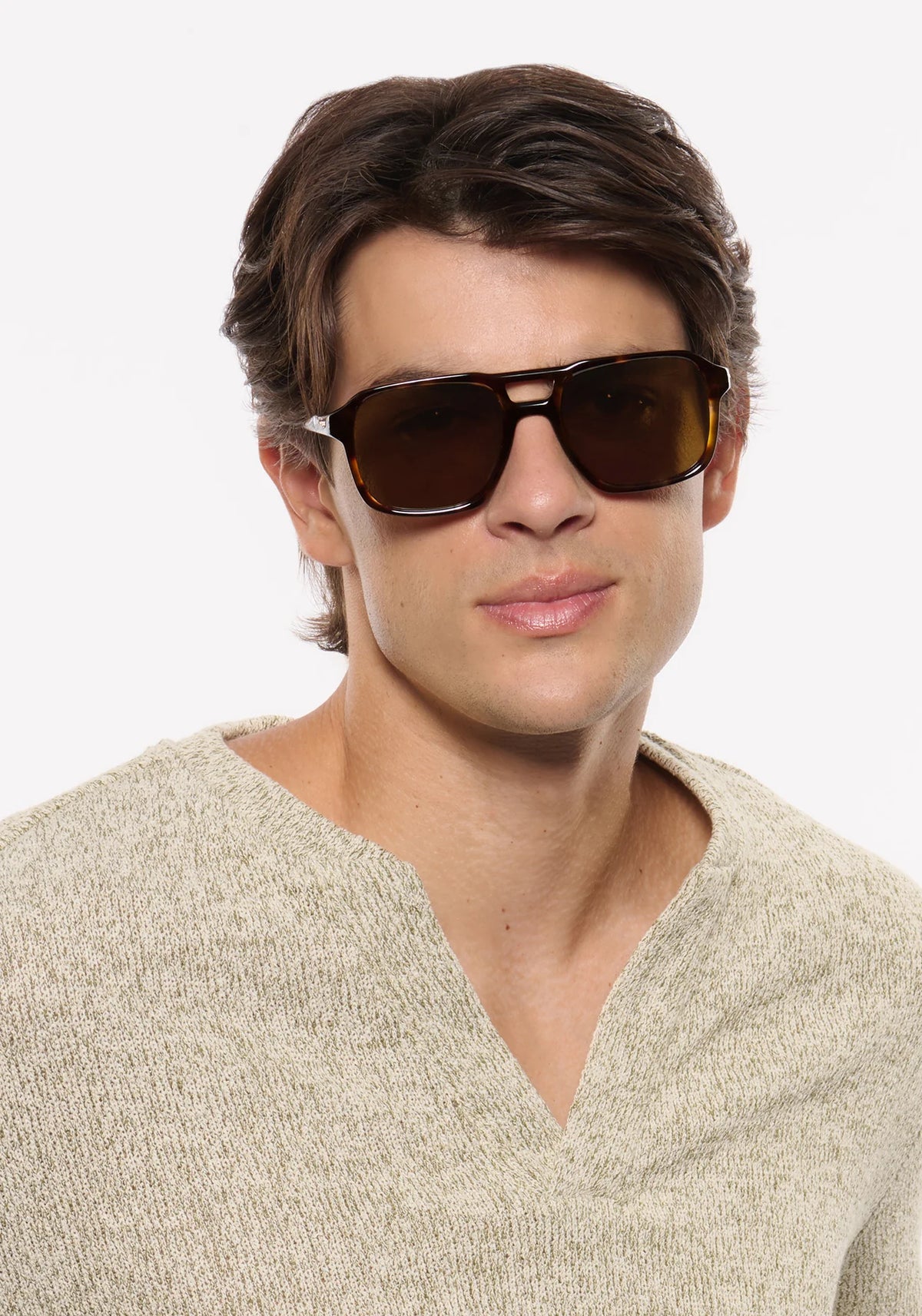 A man wearing dark brown aviator-style sunglasses with a square frame and a beige knit sweater, photographed against a clean white background.