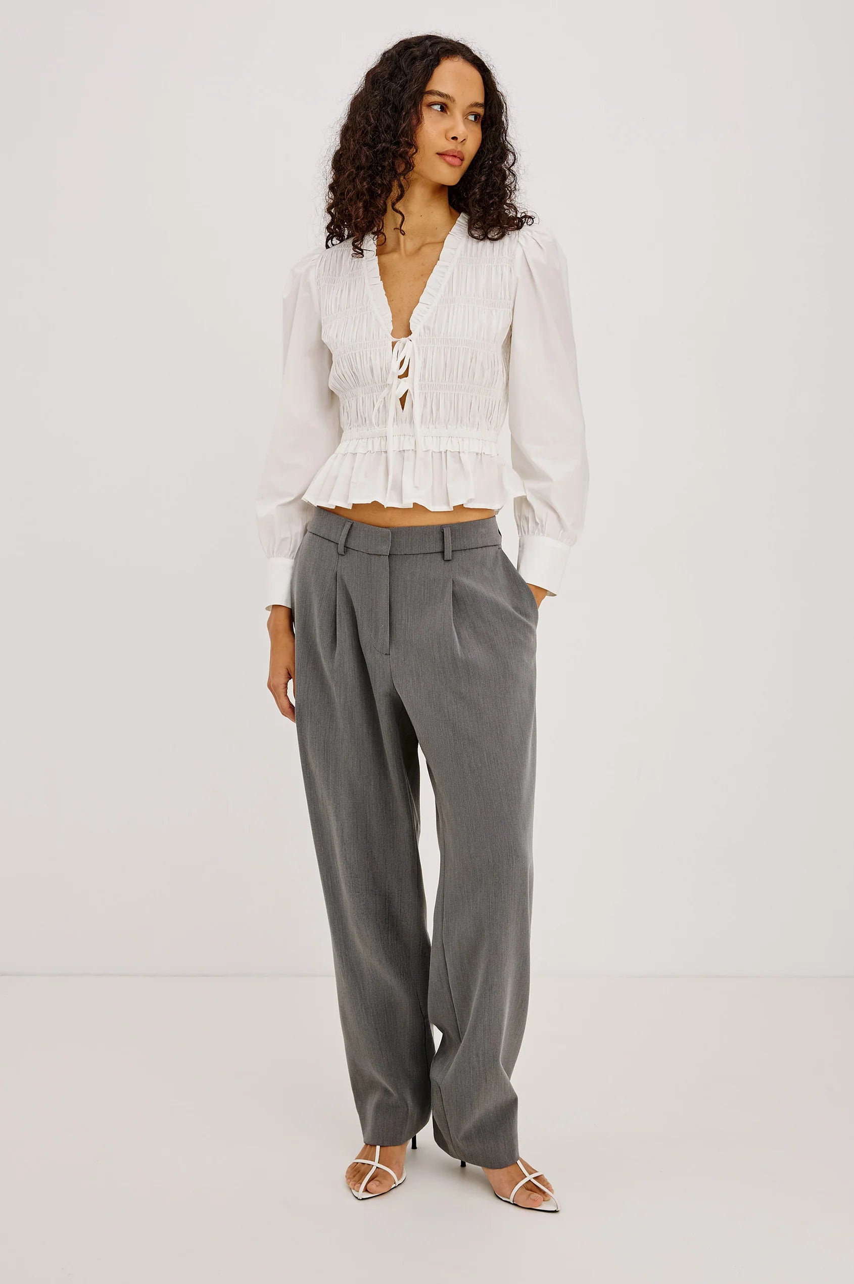 A model stands against a plain white studio background wearing a white long-sleeve crinkled blouse with a tie neckline and a gathered peplum hem, paired with high-waisted gray wide-leg trousers and strappy flat sandals.