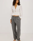 A model stands against a plain white studio background wearing a white long-sleeve crinkled blouse with a tie neckline and a gathered peplum hem, paired with high-waisted gray wide-leg trousers and strappy flat sandals.
