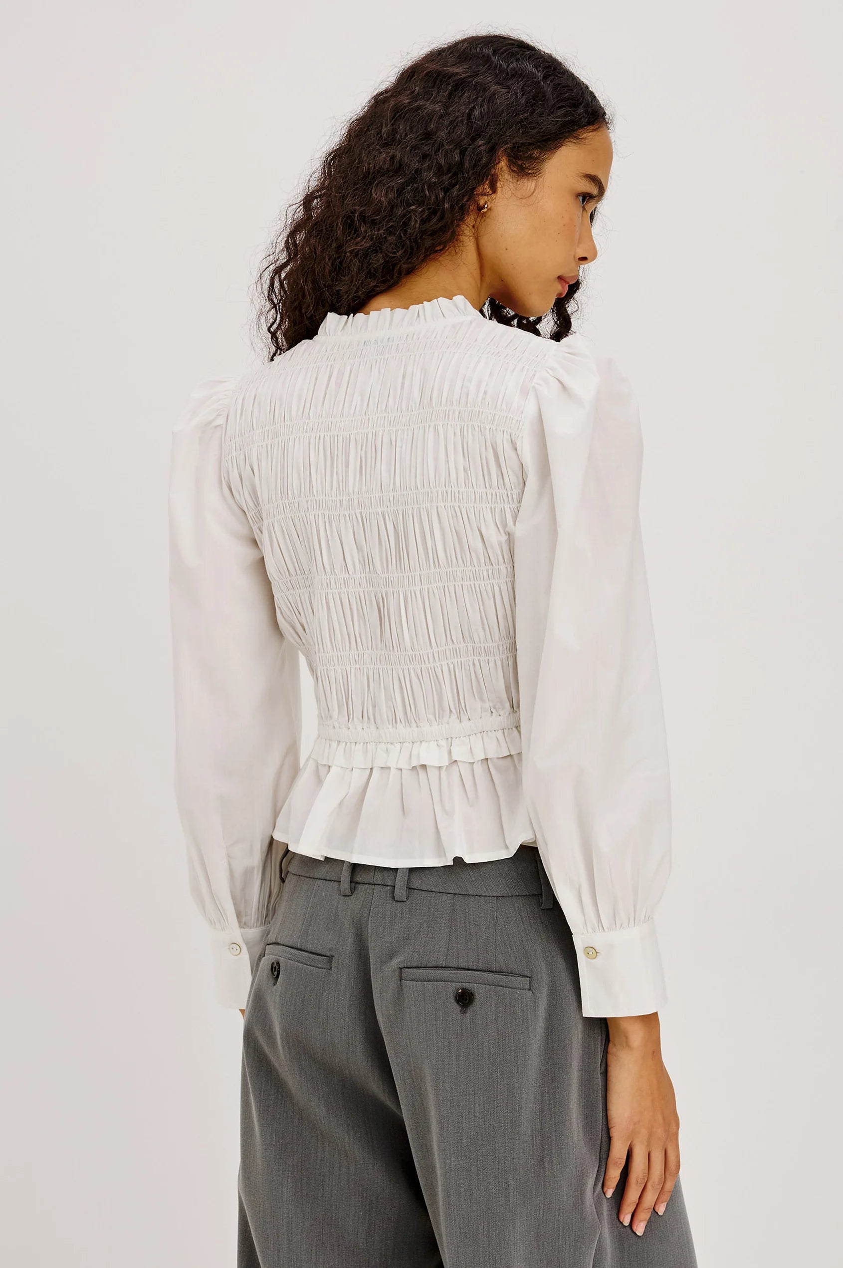 A woman shown from the back wearing a white long-sleeve blouse with a smocked back and ruffled hem, paired with gray trousers, against a plain light background.