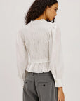 A woman shown from the back wearing a white long-sleeve blouse with a smocked back and ruffled hem, paired with gray trousers, against a plain light background.