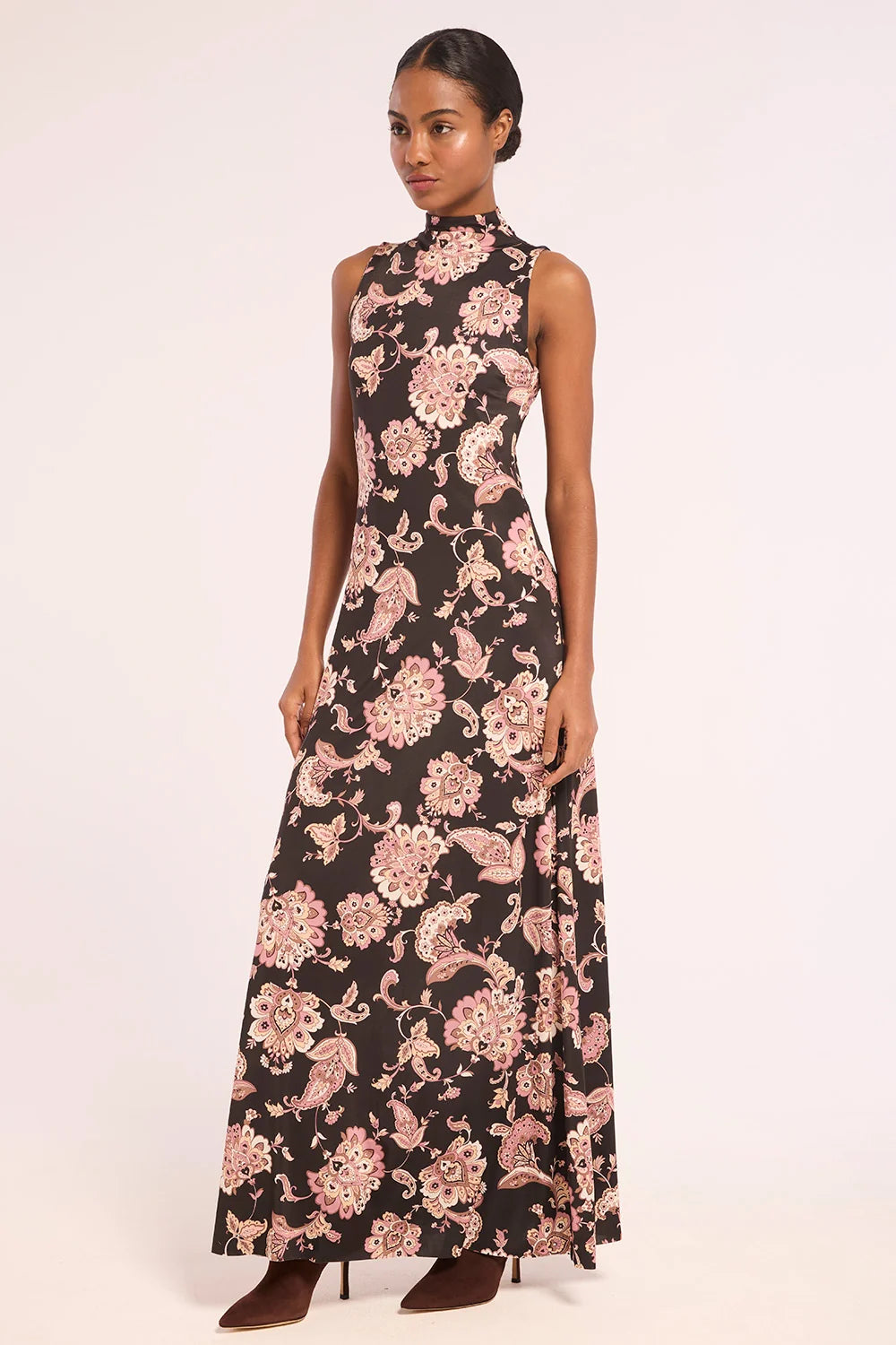 A woman modeling a sleeveless high-neck maxi dress with a black background and a pink and beige floral paisley print, standing against a plain light background.