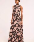A woman modeling a sleeveless high-neck maxi dress with a black background and a pink and beige floral paisley print, standing against a plain light background.