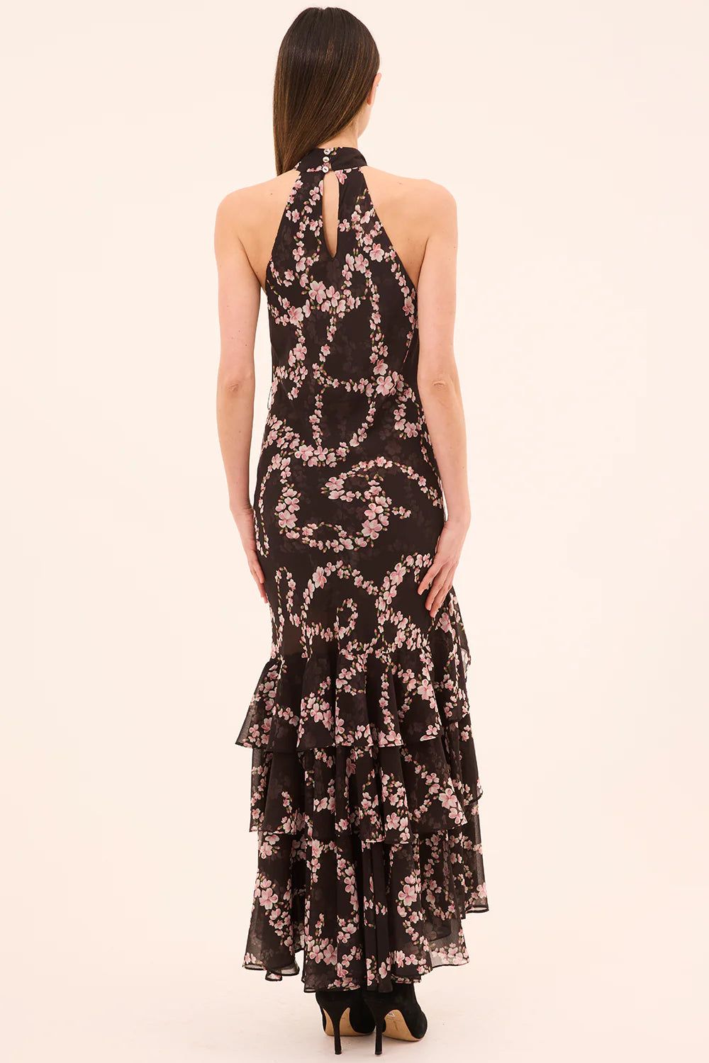 Woman wearing a black floral dress with ruffled layers on a beige background back view