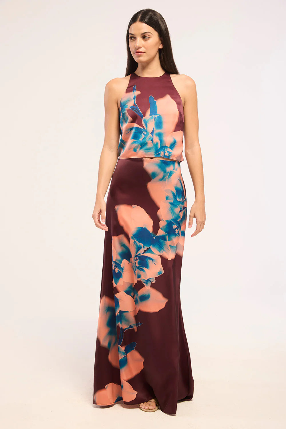 A woman modeling a sleeveless halter-neck maxi dress with a floral print in teal blue and peach-pink over a deep burgundy base, set against a plain white studio background.