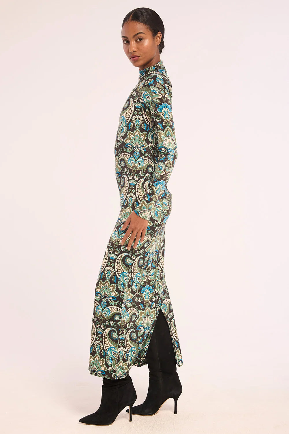 A woman modeling a long-sleeved maxi dress with a detailed paisley pattern in teal, black, and beige, paired with black ankle boots, set against a plain light background.