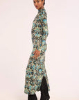 A woman modeling a long-sleeved maxi dress with a detailed paisley pattern in teal, black, and beige, paired with black ankle boots, set against a plain light background.