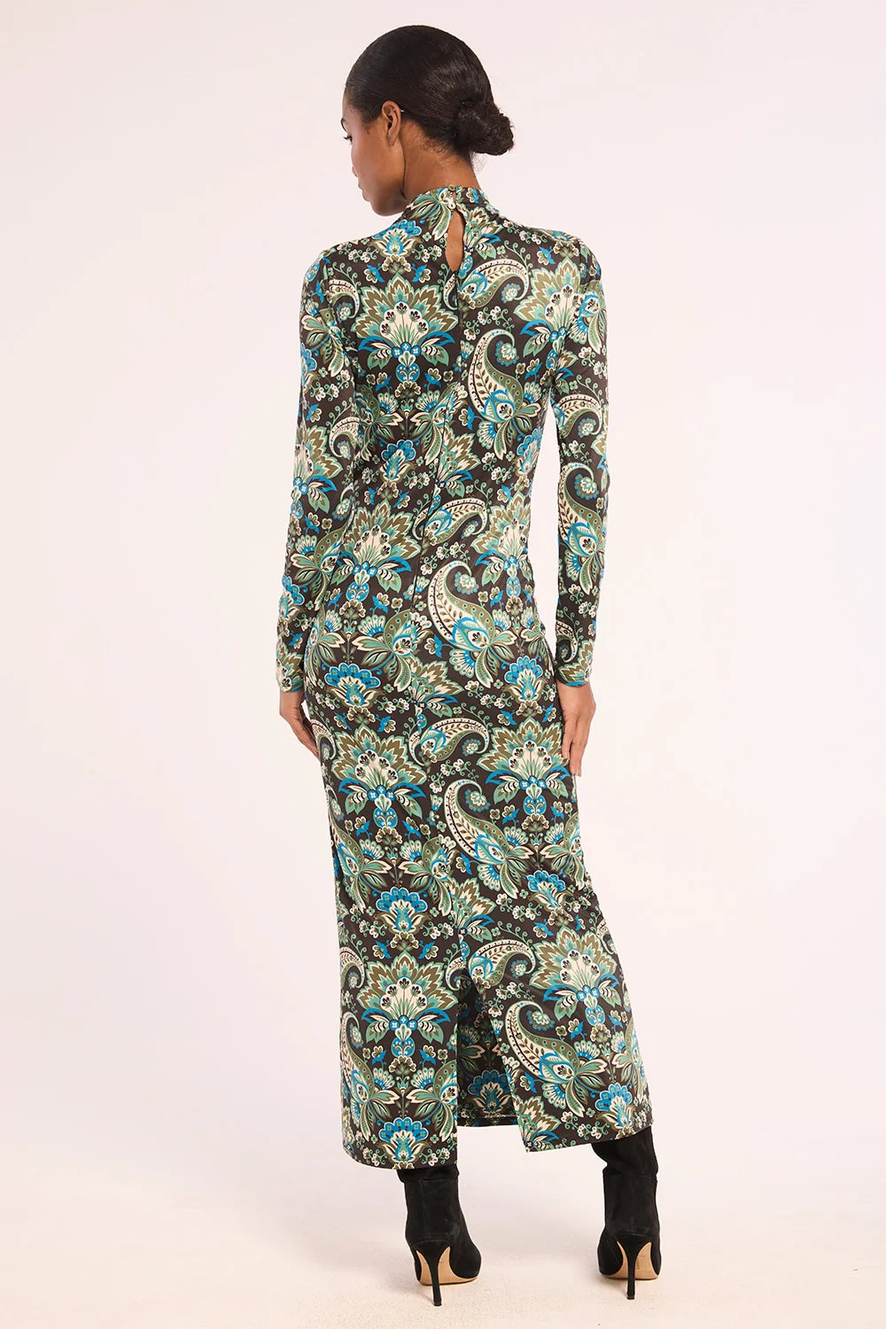 A woman shown from the back wearing a long-sleeve maxi dress with a detailed paisley pattern in teal, black, and beige, paired with black ankle boots, against a plain light background.