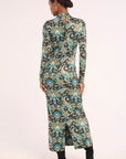 A woman shown from the back wearing a long-sleeve maxi dress with a detailed paisley pattern in teal, black, and beige, paired with black ankle boots, against a plain light background.