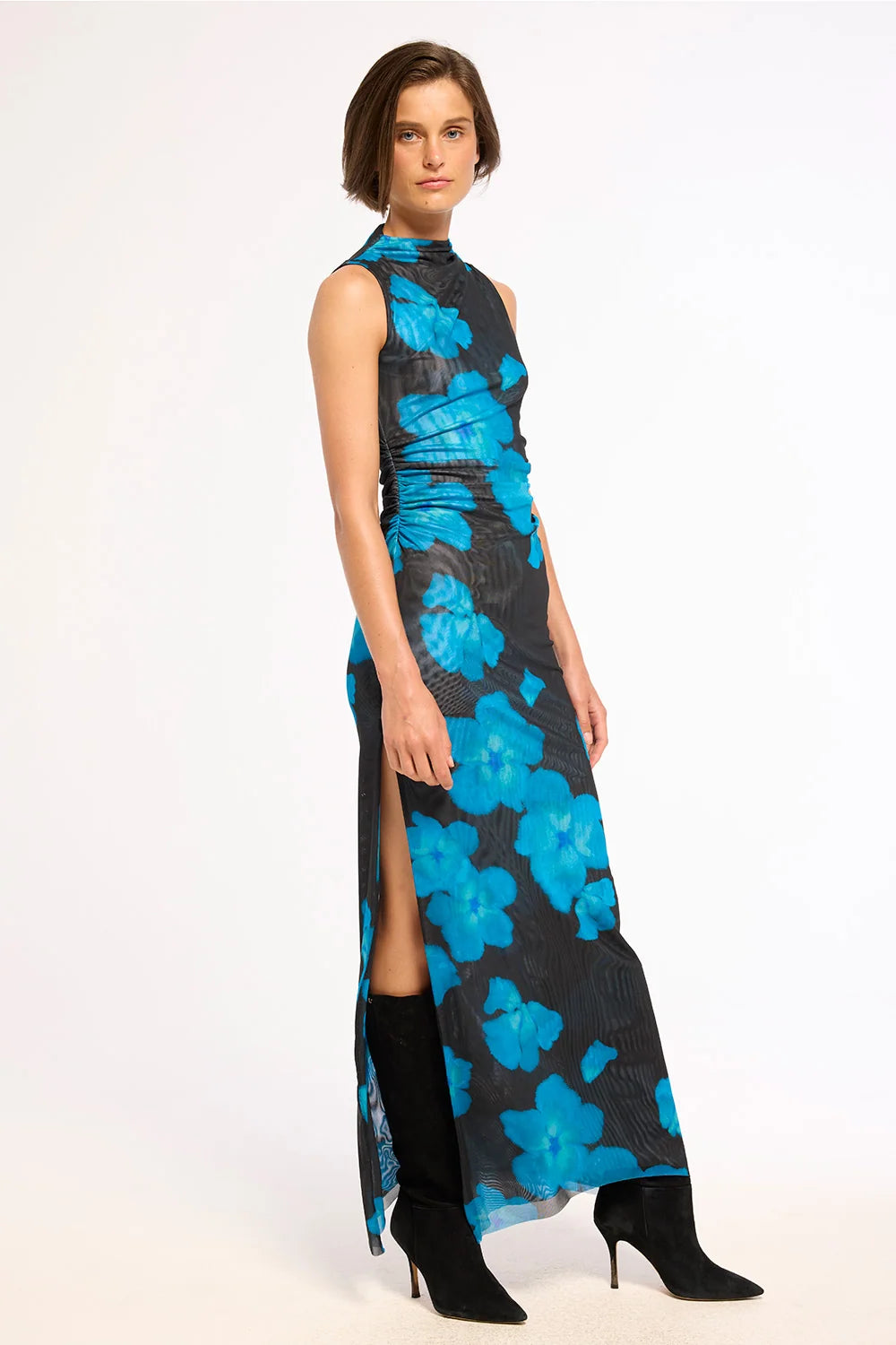 A model wearing a sleeveless, high-neck, floor-length dress with a black base and a vibrant blue floral pattern, styled with black knee-high boots against a plain white studio background.
