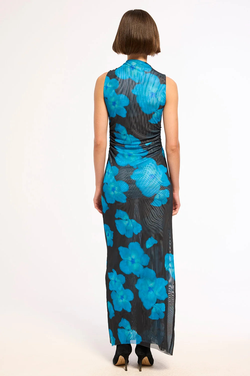 A woman viewed from the back wearing a sleeveless, floor-length dress with a black base and large bright blue floral pattern, standing against a plain white studio background.