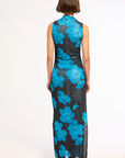 A woman viewed from the back wearing a sleeveless, floor-length dress with a black base and large bright blue floral pattern, standing against a plain white studio background.