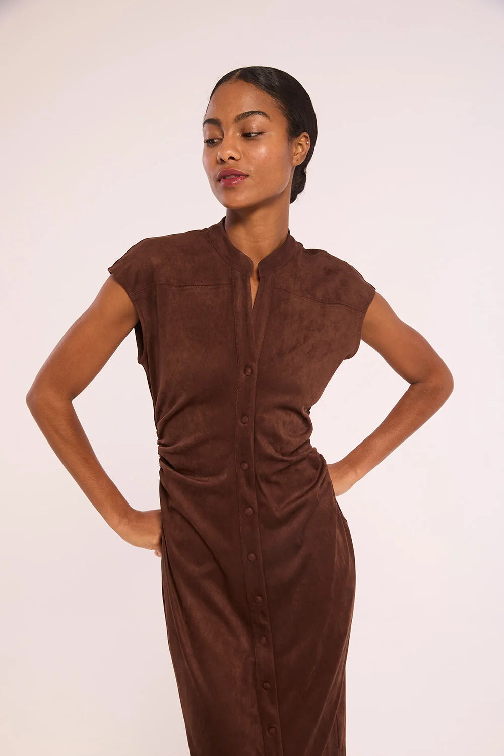 A woman modeling a sleeveless, button-front brown dress with a mock neck and gathered waist details, posed against a plain light background.
