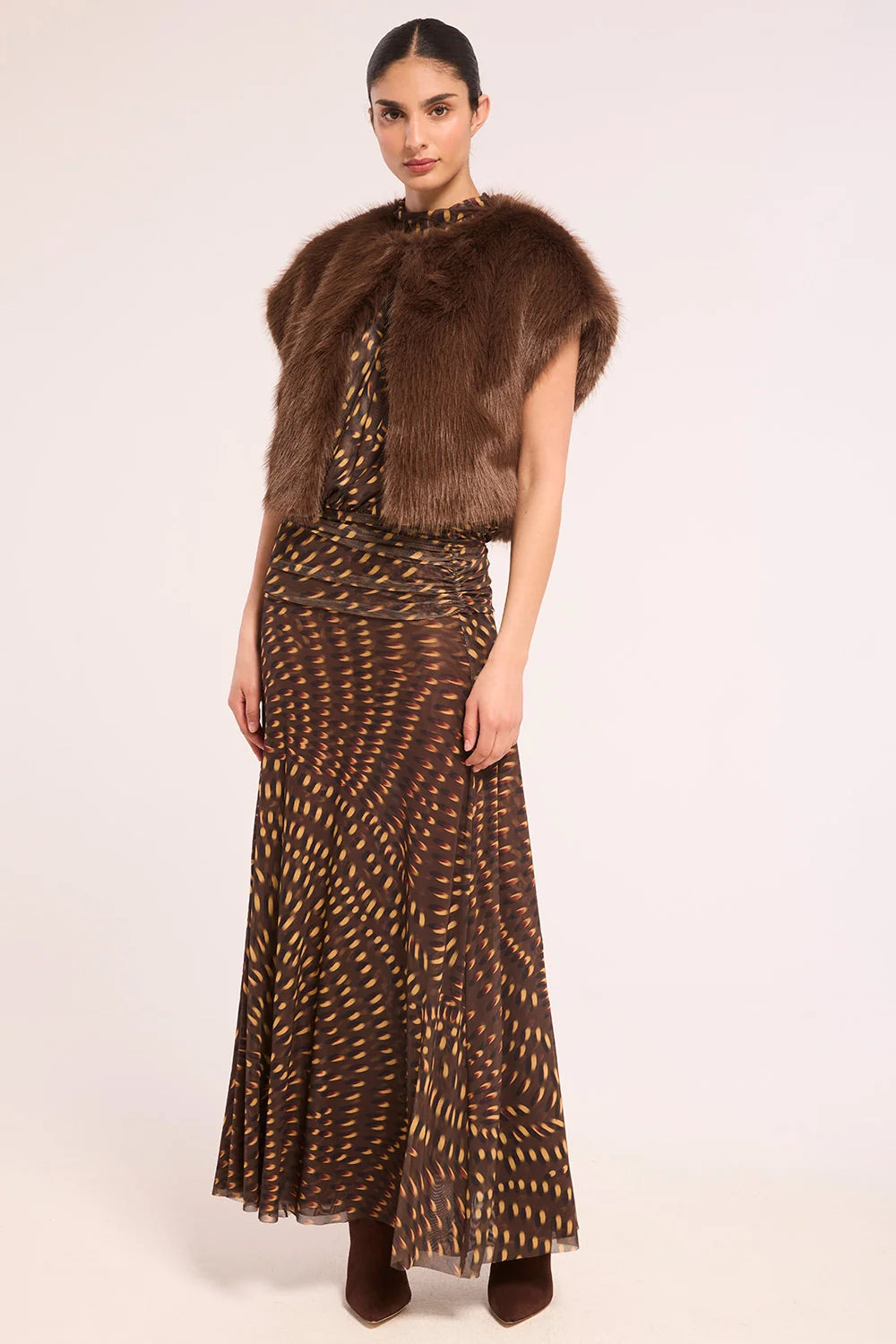 A model wearing a brown faux fur collarless vest over a brown and gold patterned dress, paired with brown pointed-toe boots, against a plain light background.
