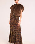 A model wearing a brown faux fur collarless vest over a brown and gold patterned dress, paired with brown pointed-toe boots, against a plain light background.