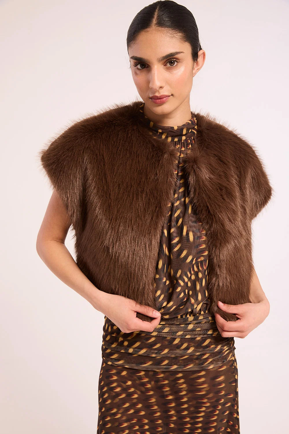 A woman modeling a brown faux fur shrug vest over a brown and gold patterned dress, set against a plain light background.