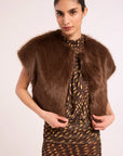 A woman modeling a brown faux fur shrug vest over a brown and gold patterned dress, set against a plain light background.