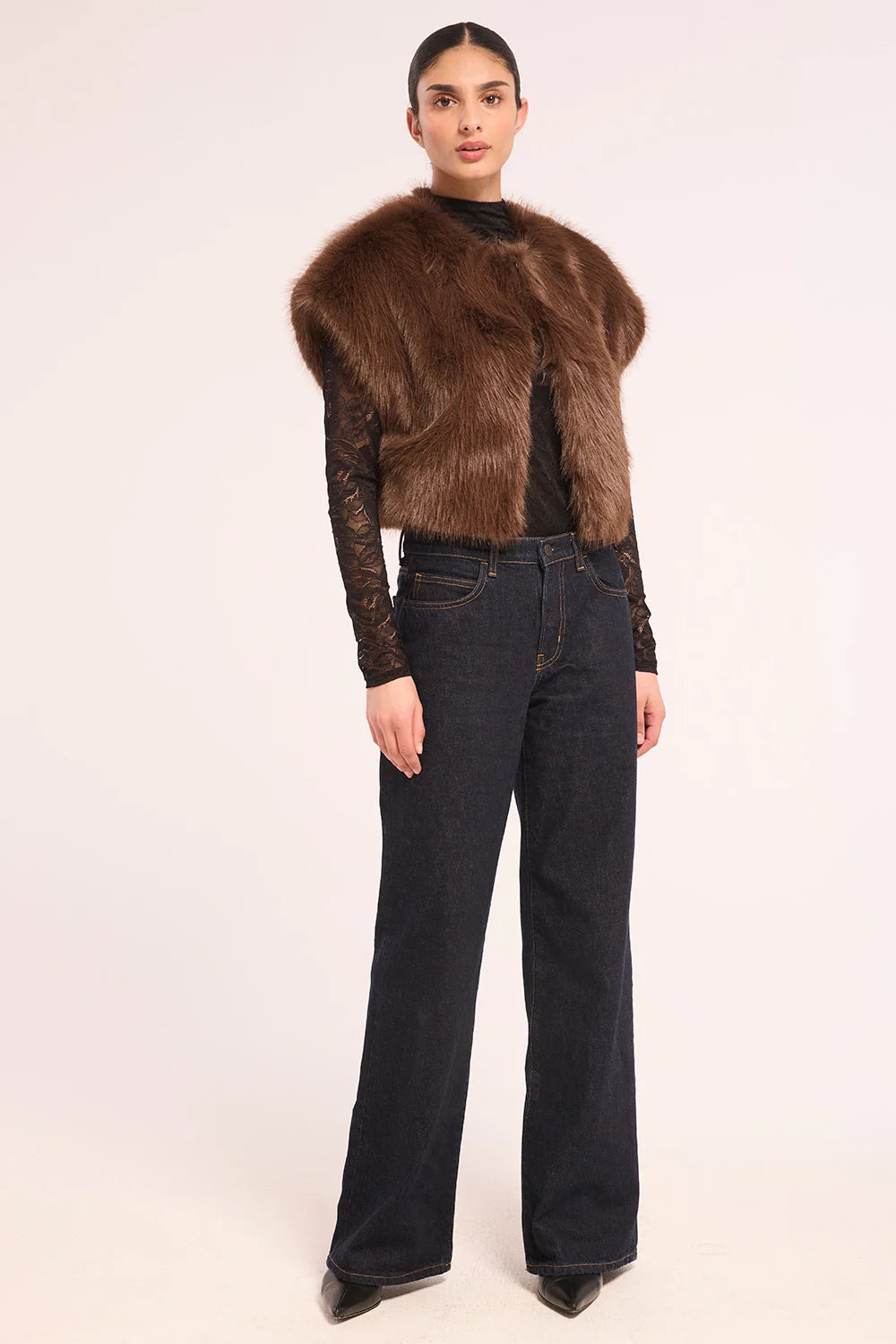 A model is shown from the back wearing a brown fur-look cropped vest over a black lace long-sleeve top and dark blue wide-leg jeans, against a plain light background.