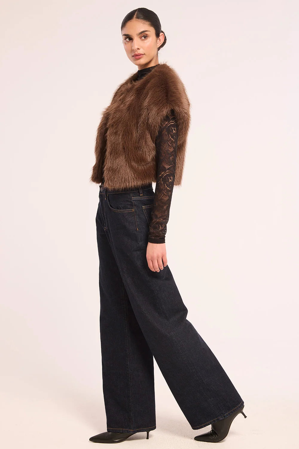 A model is shown from the side wearing a brown faux fur vest over a dark brown lace long-sleeve top, paired with dark wide-leg jeans and black pointed-toe heels, against a plain light background.