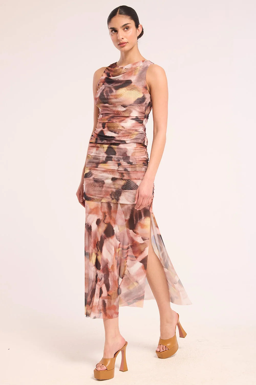 A model wearing a sleeveless, high-neck, ruched midi set with an abstract painterly print in shades of brown, beige, and pink, paired with tan platform heels, against a solid off-white background.