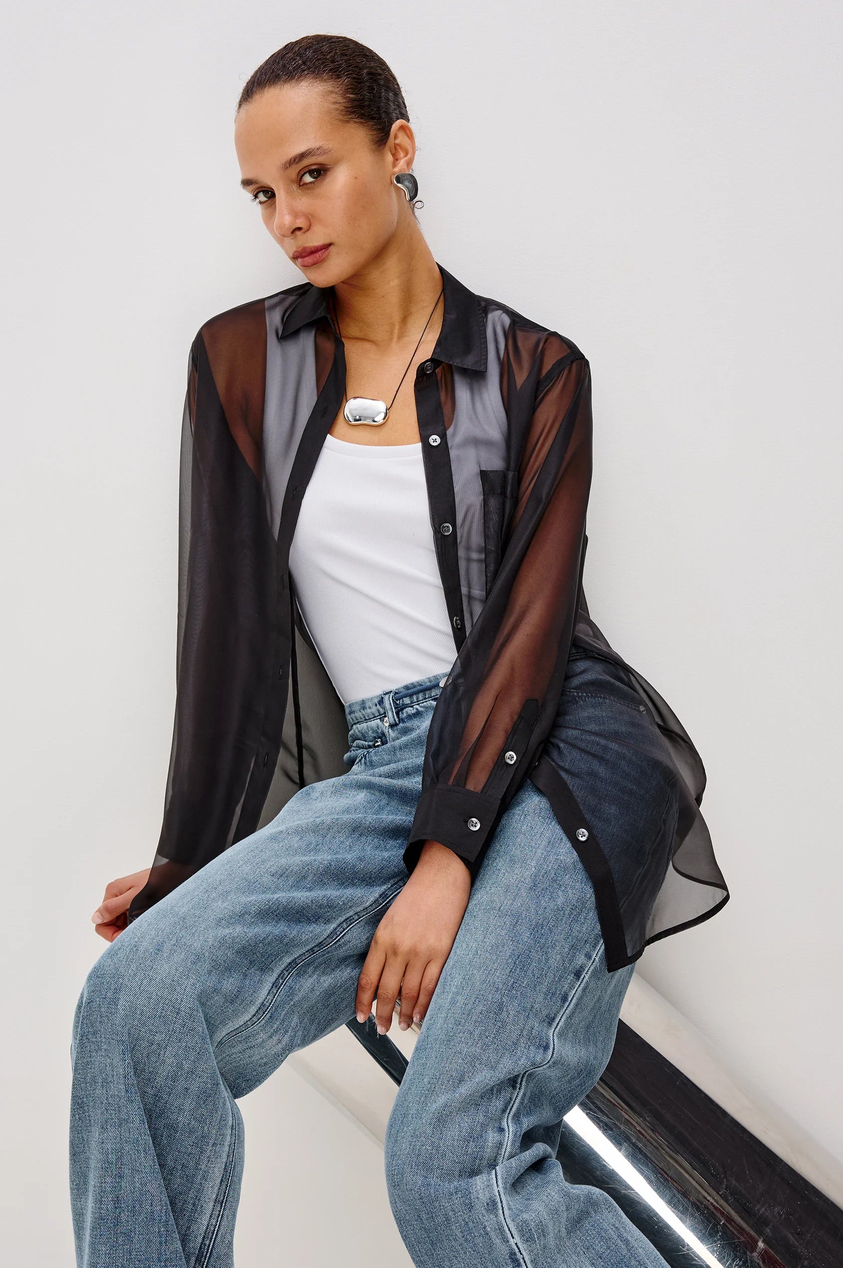 Woman wearing a sheer black shirt over a white top and blue jeans on a light gray background