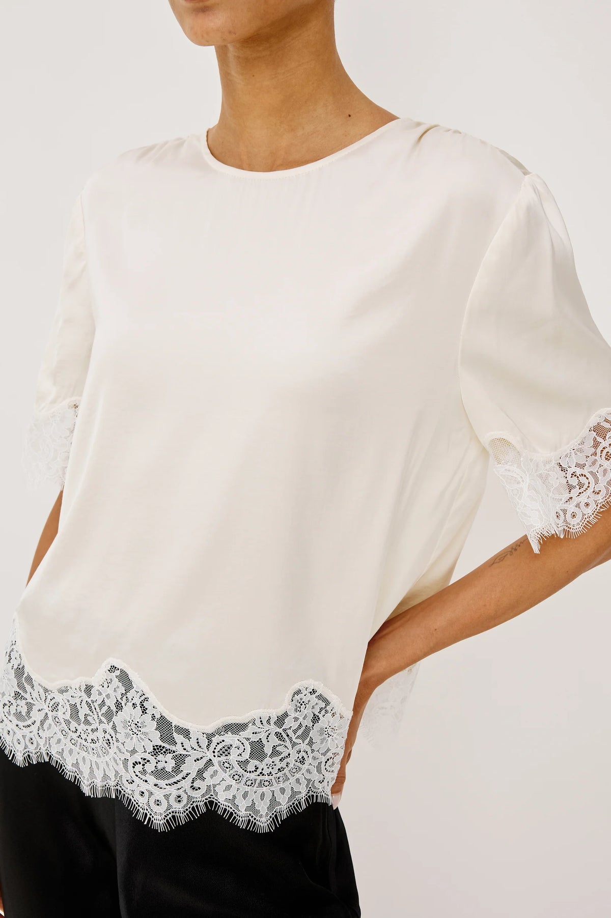 An off-white short-sleeve blouse with delicate white lace trim at the hem and sleeve cuffs, worn by a model against a light gray studio background.
